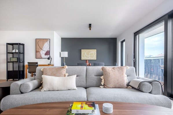 A modern living room with a grey sofa, a coffee table with books, a bookshelf, and a city view through large windows.