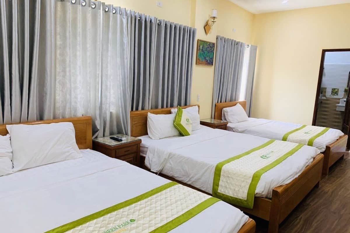 A hotel room with four beds, each with white bedding and green accents, wooden headboards, and a large window with curtains.