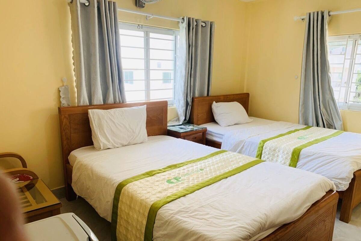 A hotel room with two single beds, a wooden headboard, a bedside table, a chair, and a window with curtains.