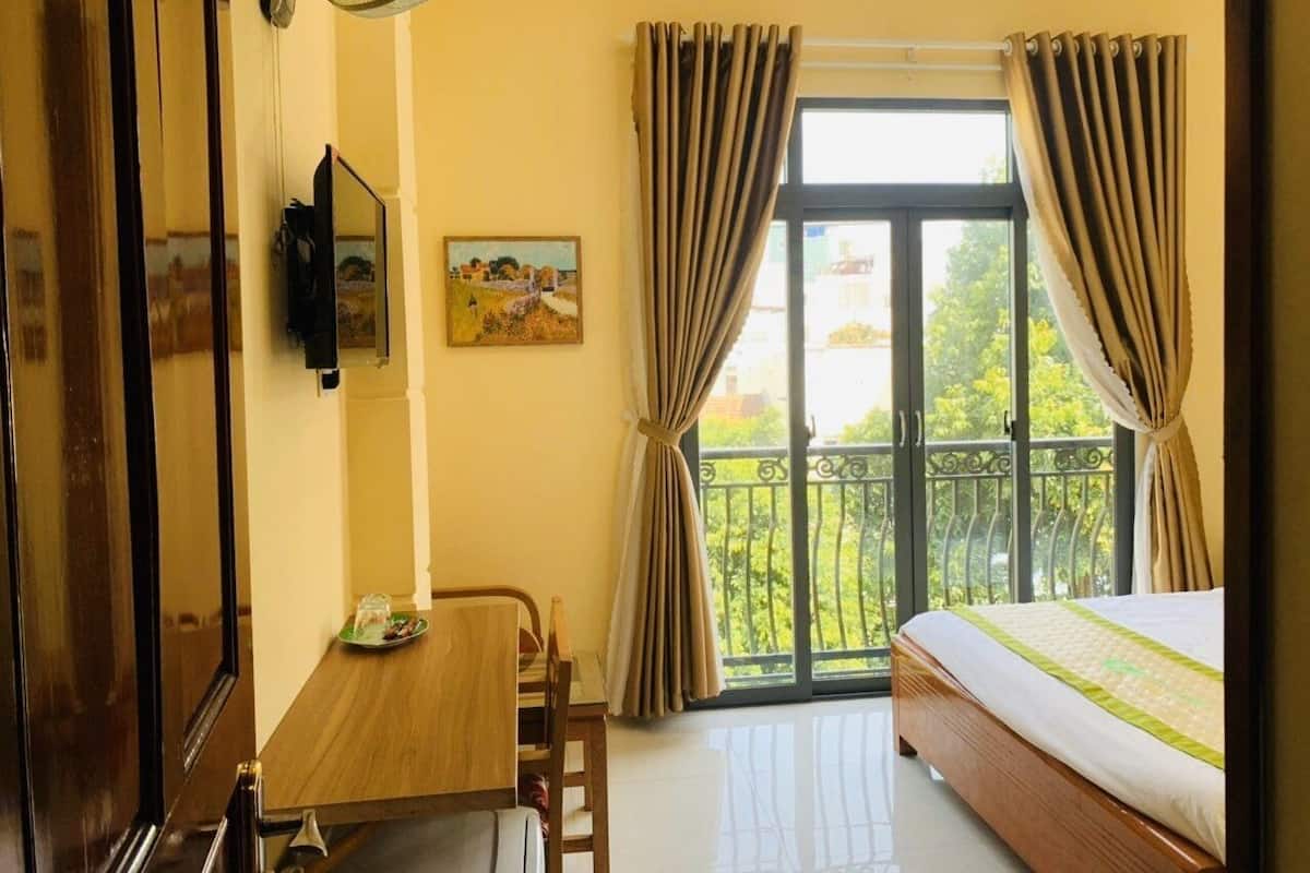 A hotel room with a bed, a dining table, a chair, a TV, a painting, and a balcony with curtains.