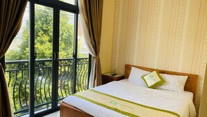 A hotel room with a large bed, a wooden headboard, a nightstand, and a view of greenery through a balcony door. - Green Park 2 Hotel (Quy Nhon Nam)