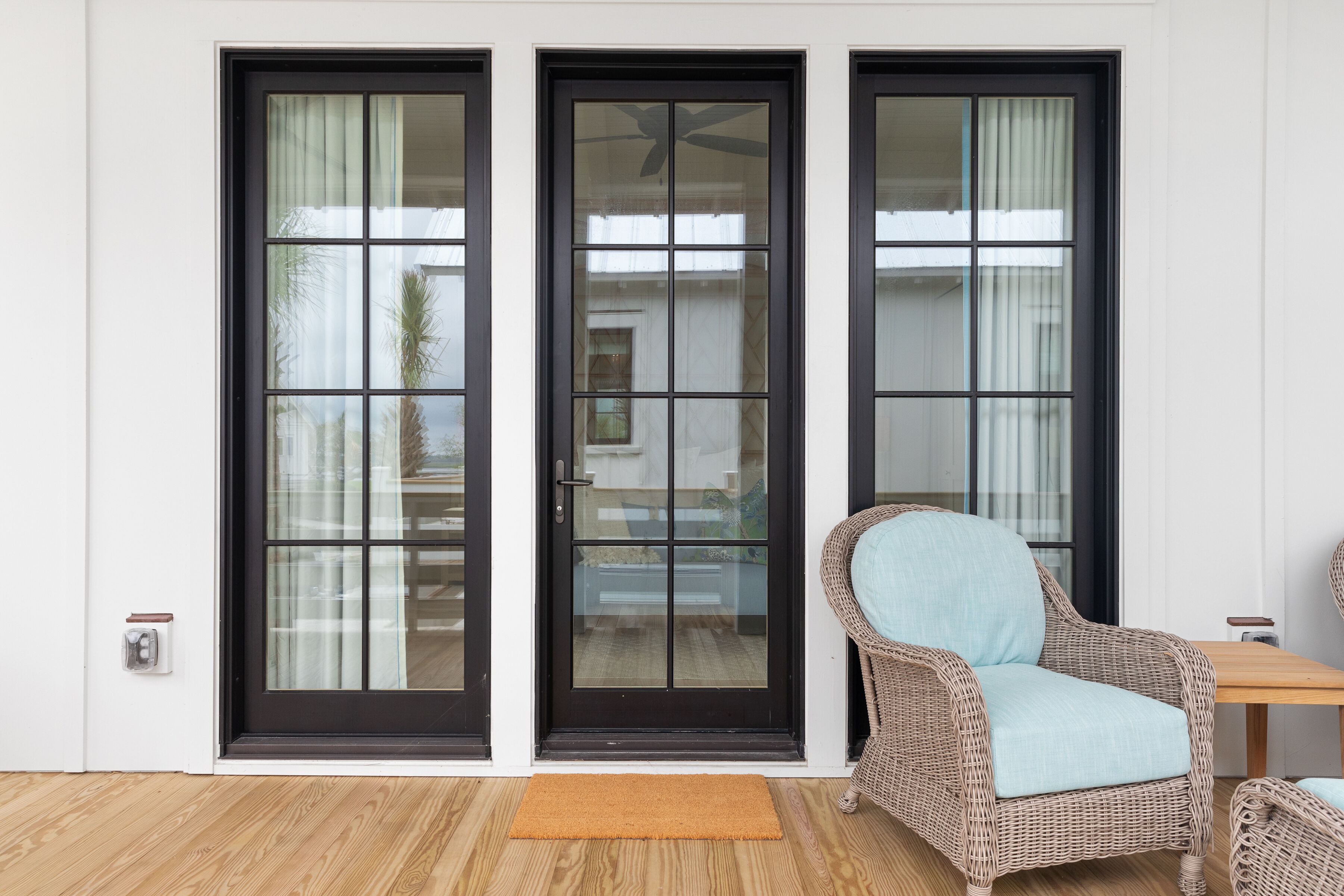 Una habitación con tres puertas de vidrio con marco negro, una silla de mimbre con cojín azul claro y una mesa de madera.
