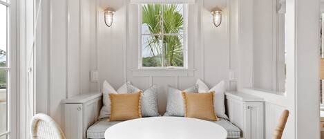 A white room with a round table, a bench with cushions, and a window with a plant.