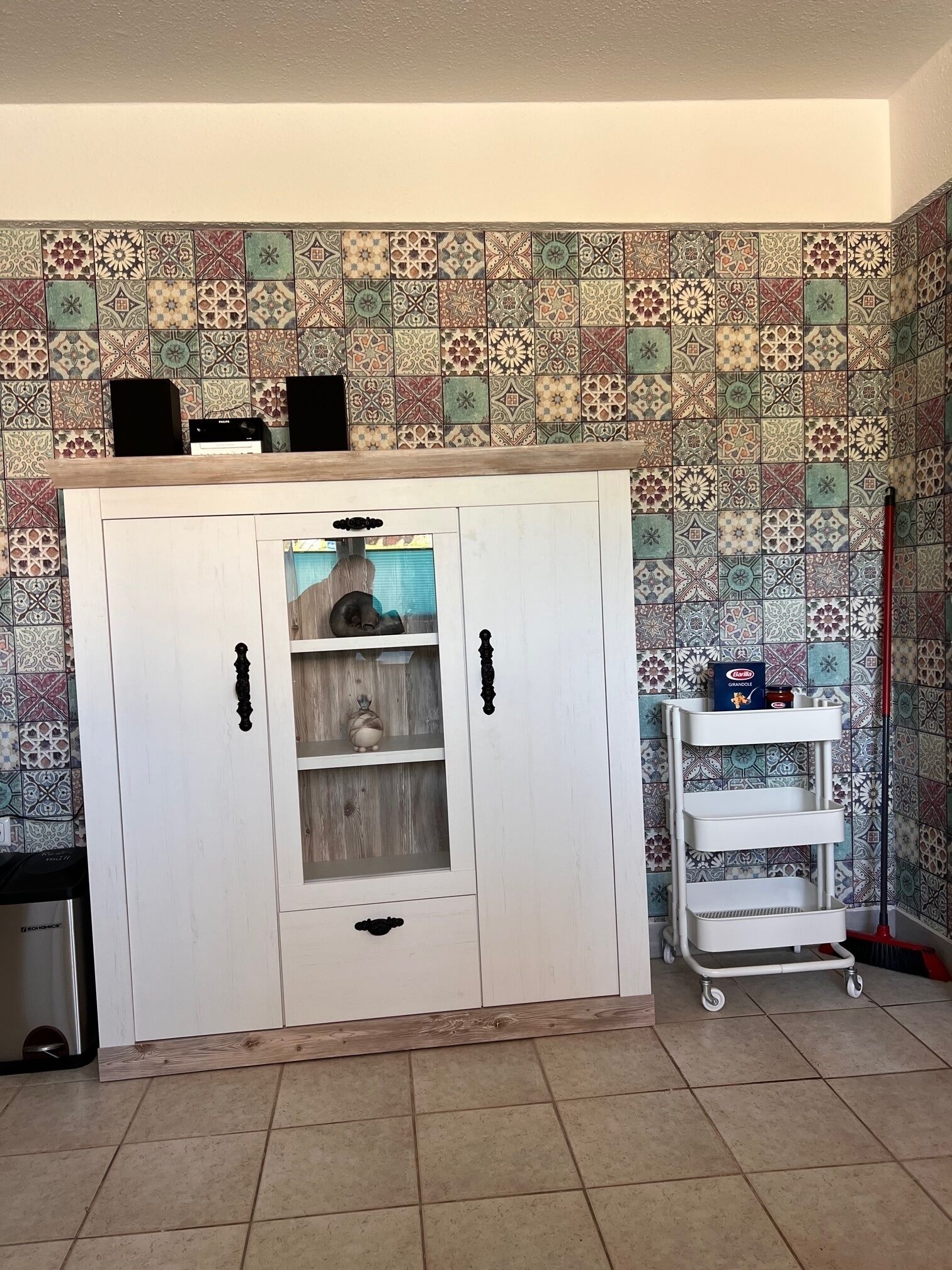 A white cabinet with multiple shelves and a glass door, a white shelf unit with a box labeled 'GURANDELLE,' and a broom leaning against the wall.
