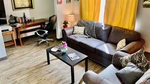 A living room with a sofa, coffee table, and a desk with a chair. There are curtains, a lamp, and artwork on the walls.