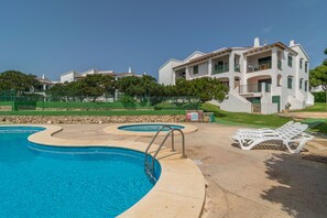 A swimming pool with a curved design, a set of white sun loungers, and a two-story building with balconies.