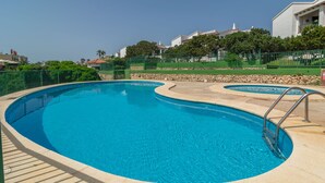A swimming pool with a curved design, surrounded by a stone wall and a green fence. - BINIFORCAT 11 - Charming apartment with shared pool located 250 meters from the beach. (Ciutadella de Menorca)