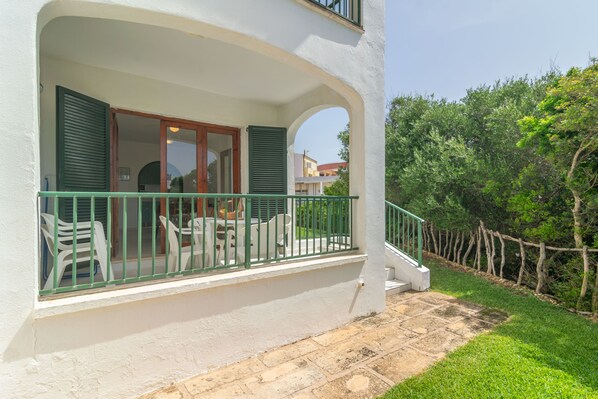A white building with a balcony featuring a green railing, outdoor seating, and a wooden staircase leading to the entrance.