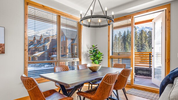 A dining area with a wooden table and chairs, a chandelier, and large windows with blinds. - Spring Creek Vacations 2 BR Premier Condo 107A (Canmore)