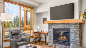 A modern living room with a stone fireplace, comfortable seating, a flat-screen TV, and large windows with blinds. - Spring Creek Vacations 2 BR Premier Condo 107A (Canmore)