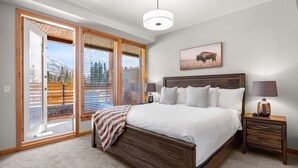 A bedroom with a large bed, wooden headboard, a nightstand with a lamp, and a window with blinds. - Spring Creek Vacations 2 BR Premier Condo 107A (Canmore)