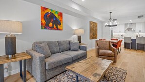Un salon moderne avec un canapé gris, une table basse en bois et un tapis à motifs. Un tableau abstrait aux couleurs vives est accroché au mur.