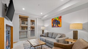 Un salon moderne avec une cheminée, un canapé gris, un fauteuil marron, une table basse et un tableau abstrait aux couleurs vives accroché au mur.