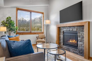 A living room with a fireplace, a sofa with cushions, a chair, a table, and a large window with blinds. - Spring Creek Vacations 1 BR Elevated Condo 403A (Canmore)