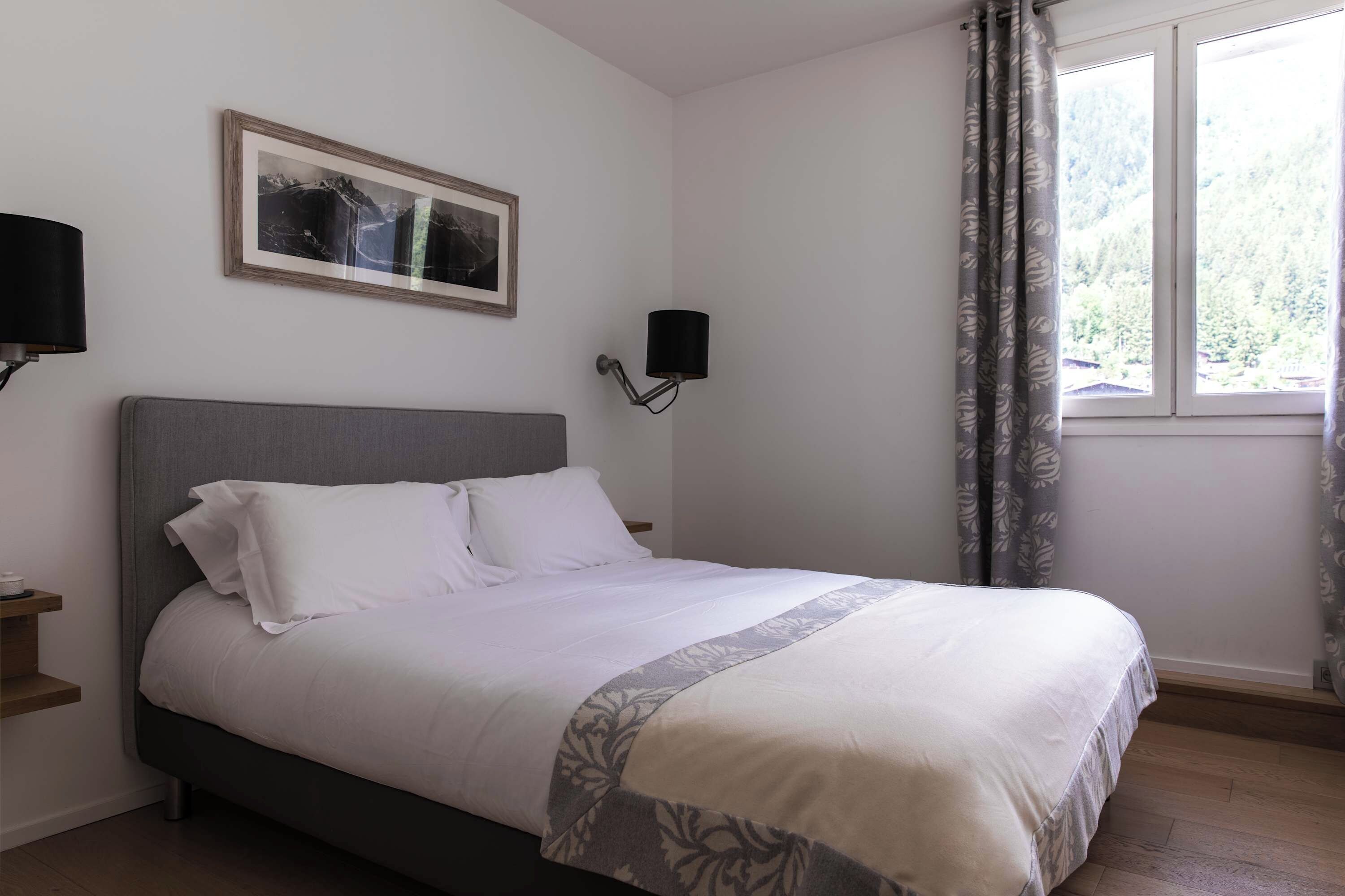 A bedroom with a bed, a nightstand, a wall-mounted lamp, a framed picture, and a window with curtains.