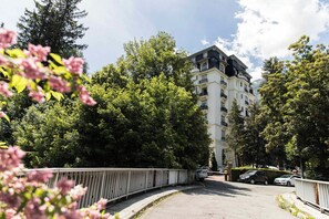 A multi-story building with a white facade and black accents, surrounded by lush greenery and a pedestrian bridge. - Majestic 145 Historical Building Chamonix All Year (Chamonix)