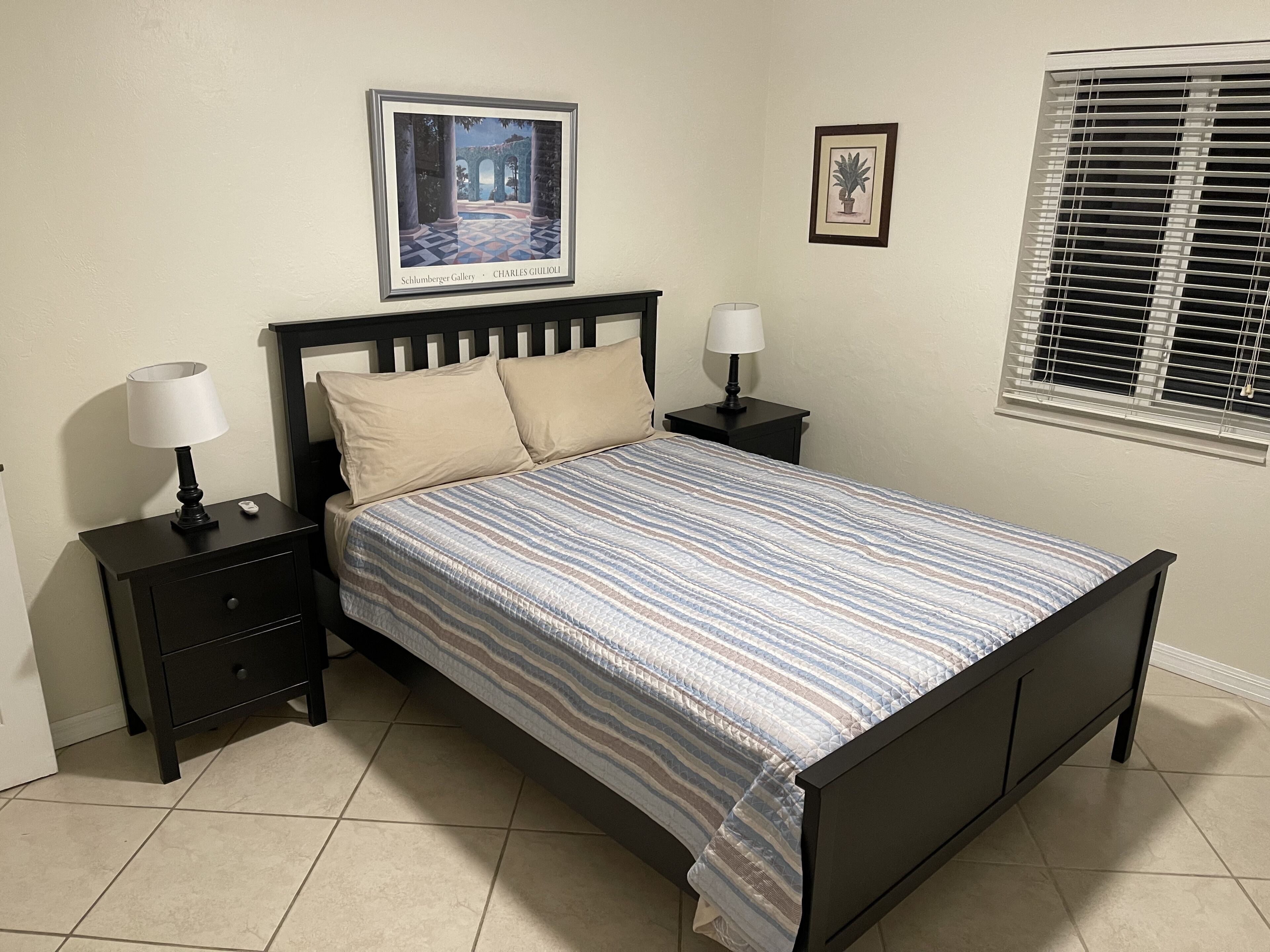 Un dormitorio con una cama, dos mesitas de noche, una lámpara de noche, un cuadro enmarcado en la pared y una ventana con persianas.