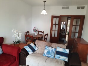 A living room with a beige sofa, red armchair, and a wooden dining table with chairs. There is a vase with flowers on the table and a ceiling light fixture. - Central 2-Bed Apartment in Estepona Port (Estepona)