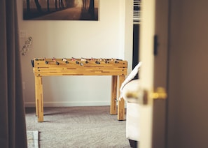 Una mesa de fútbol de madera en una habitación con piso alfombrado, un cuadro en la pared y una puerta al frente.