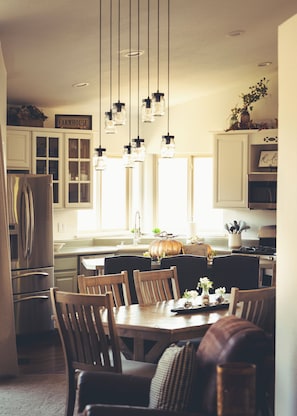 A kitchen with white cabinets, a dining table with chairs, hanging pendant lights, and a refrigerator.