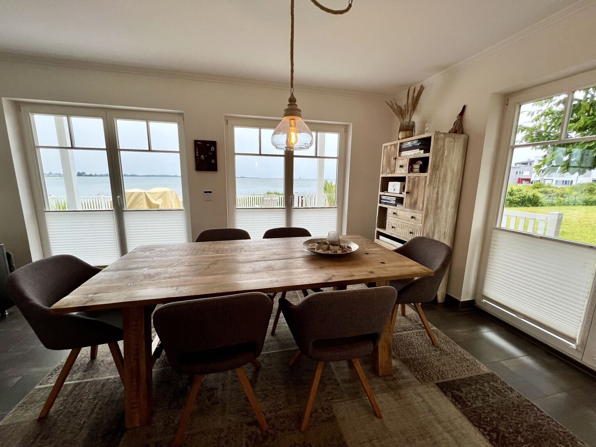 A dining area with a wooden table, six chairs, a hanging light fixture, and large windows with a view of the sea.