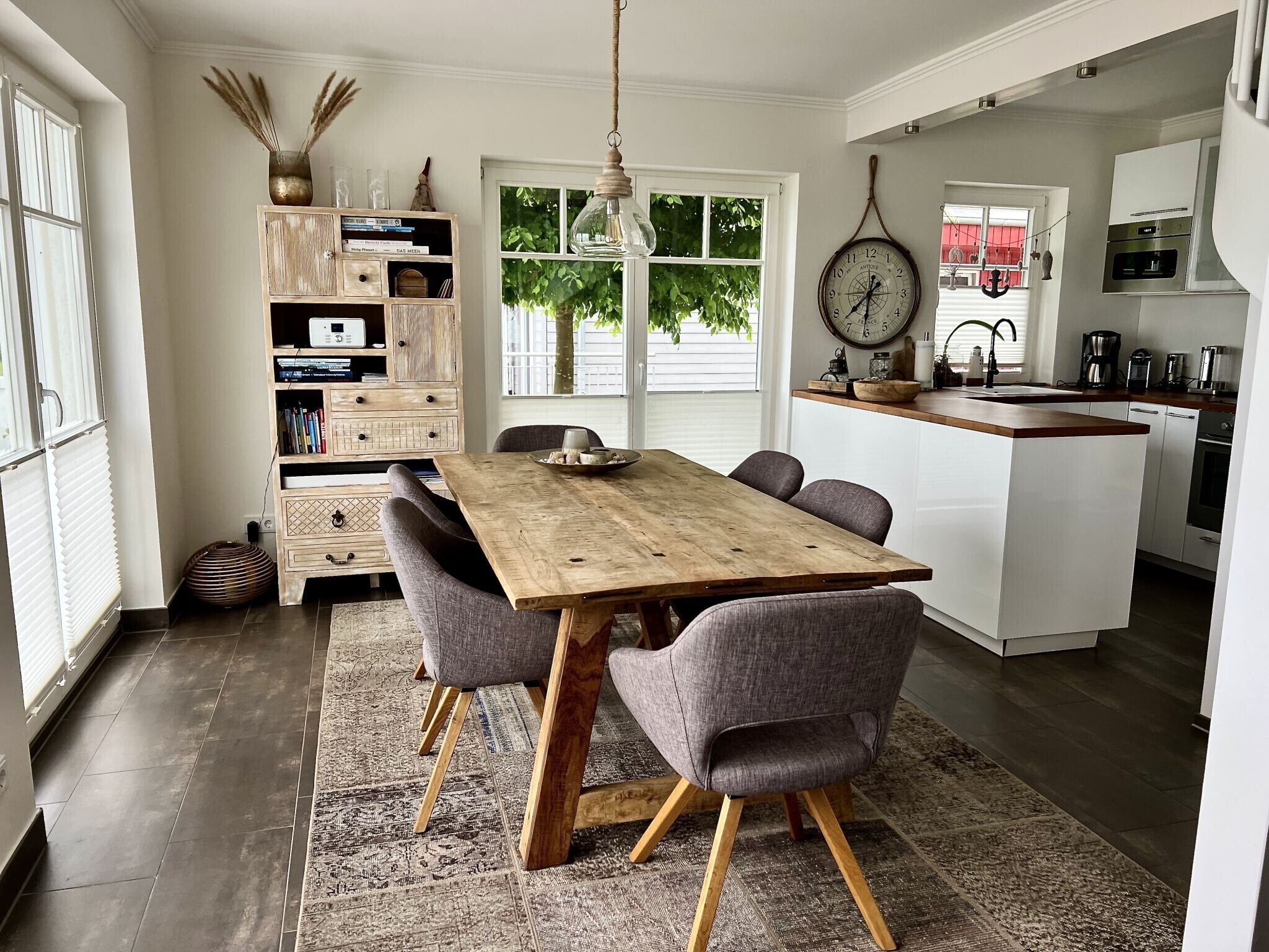 Un coin repas avec une table en bois et des chaises à revêtement gris, une cuisine avec des meubles blancs et des appareils modernes, et une grande fenêtre donnant sur un espace verdoyant.