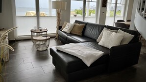 Ein modernes Wohnzimmer mit einem schwarzen Ledersofa, einem runden Holztisch und großen Fenstern mit Meerblick.