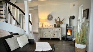 Ein Wohnzimmer mit einer schwarzen Ledersofa, einer weißen Tagesdecke, einem Holzschrank mit der Aufschrift 'BEACH' und einem Kamin, sowie einer Treppe.
