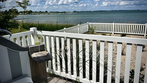 Eine Holzterrasse mit weißen Geländern, mit Blick auf ein Gewässer, eine entfernte Küste und einen klaren Himmel.