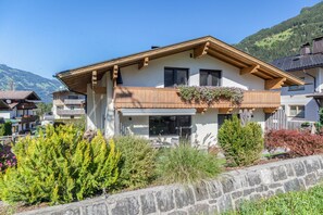 A two-story chalet with a balcony, surrounded by lush greenery and a stone wall.