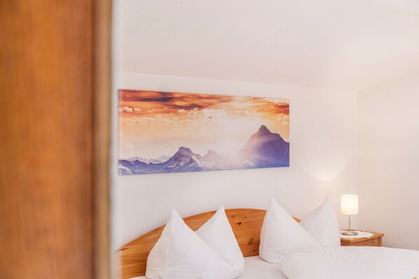 A hotel room with a bed, pillows, a nightstand, a lamp, and a wall with a mountain landscape painting.
