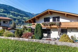 A two-story house with a balcony, surrounded by greenery and a mountain view.