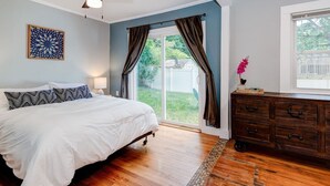 A bedroom with a bed, a ceiling fan, a window with curtains, a dresser, and a view of the backyard.