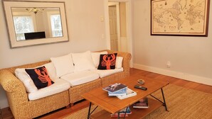Ein Wohnzimmer mit einem weißen Sofa, einem gewebten Sessel, einem Couchtisch aus Holz mit Büchern, einer gerahmten Weltkarte und einem Spiegel an der Wand.