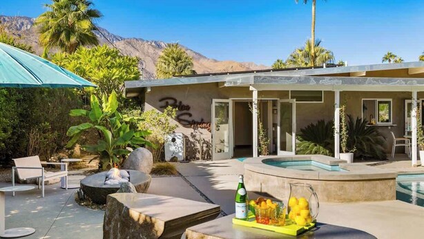 A pool area with a lounge chair, a table with a bottle and fruit, and a building with a sign. - Sunset Glamour - Enchanting Luxury Zen Retreat (Palm Springs)