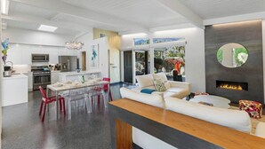 A modern interior with a kitchen, dining area, and living space, featuring a fireplace, a large mirror, and a view of the outdoors. - Sunset Glamour - Enchanting Luxury Zen Retreat (Palm Springs)