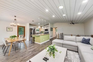 A modern living room with a sofa, a dining table with chairs, a kitchen area, and a ceiling fan.