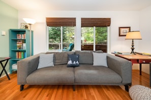 Relax in this inviting living room with a sofa, bookshelf, and desk, surrounded by large windows and wooden flooring.