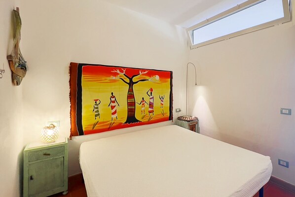 A bedroom with a bed, a nightstand, a wall-mounted lamp, a decorative wall hanging, and a window.