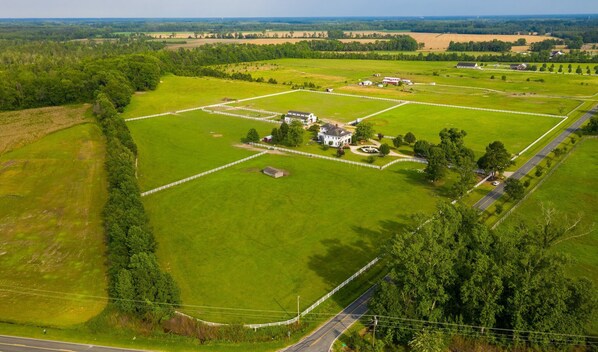 Vista aérea de una propiedad grande y rectangular con un edificio central blanco, rodeado de campos verdes y una zona cercada.