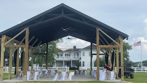 A covered outdoor event space with tables set up for an event, a large house in the background, and an American flag on a flagpole. - Parkton Place Campsite #4 on farm with pond! (Parkton)