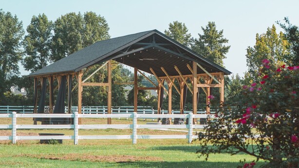 A wooden pavilion with a sloped roof, surrounded by a white fence, set in a grassy field with trees in the background. - Parkton Place Campsite #4 on farm with pond! (Parkton)