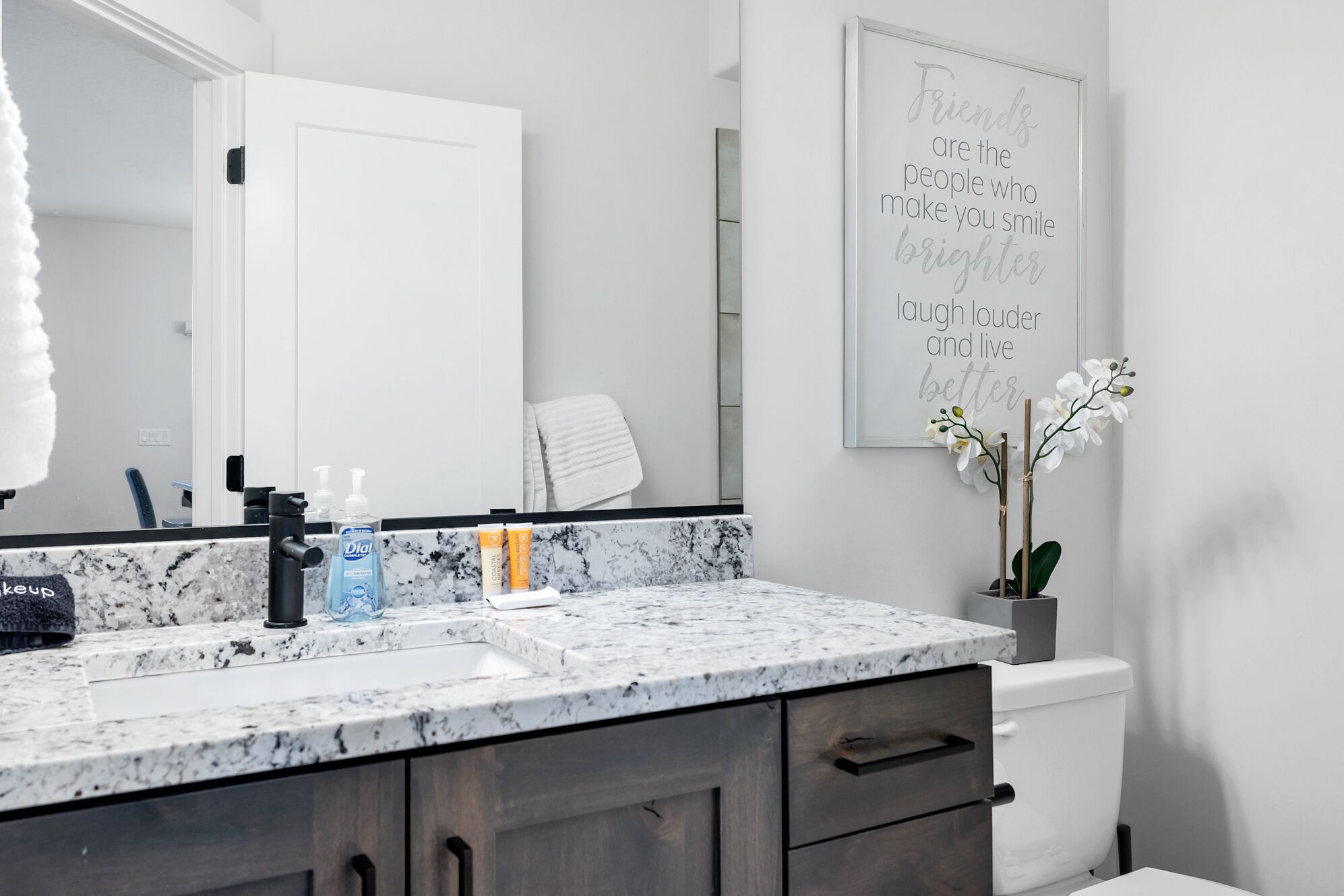 A bathroom with a marble countertop, a sink, a mirror, a toilet, and a wall-mounted sign with a motivational quote.