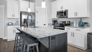 A modern kitchen with white cabinetry, stainless steel appliances, a granite countertop, and bar stools. - New Townhouse St George area, VidaSol (Washington)