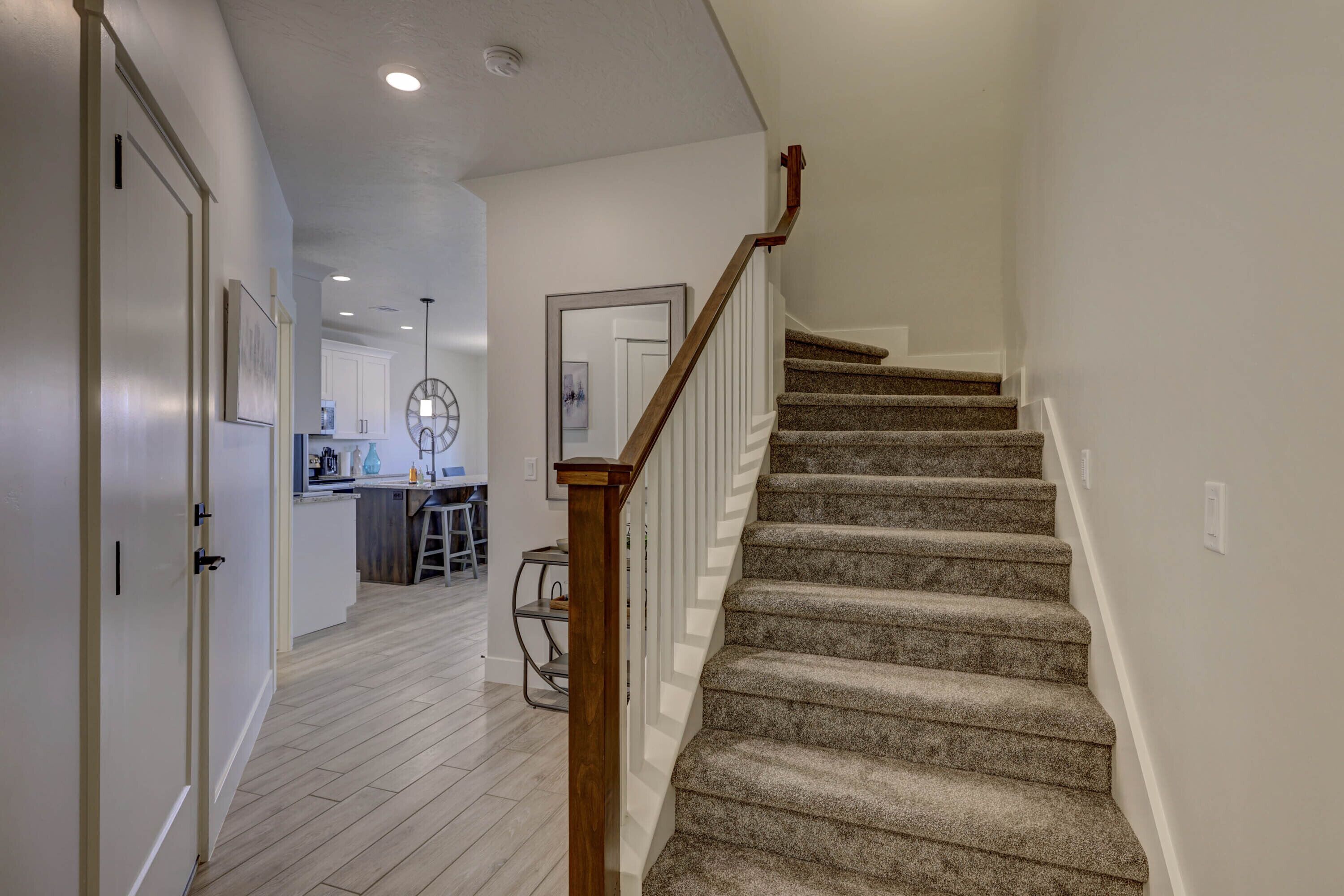 Un escalier recouvert de moquette, doté de rampes en bois, mène à un palier. On aperçoit à l’arrière-plan un coin cuisine avec des meubles blancs et une table à manger.