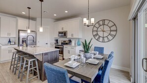 A modern kitchen with white cabinets, a central island with stools, stainless steel appliances, and a large wall clock. - New Townhouse St George area, VidaSol (Washington)