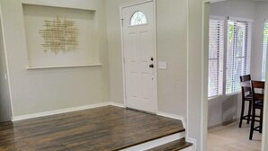 Una habitación con piso de madera, una puerta blanca con panel de vidrio y una escalera que lleva a otra zona.