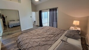 Un dormitorio con una cama grande, un ventilador de techo, una mesita de noche con lámpara, una ventana con cortinas y un espejo que refleja parte de la habitación.