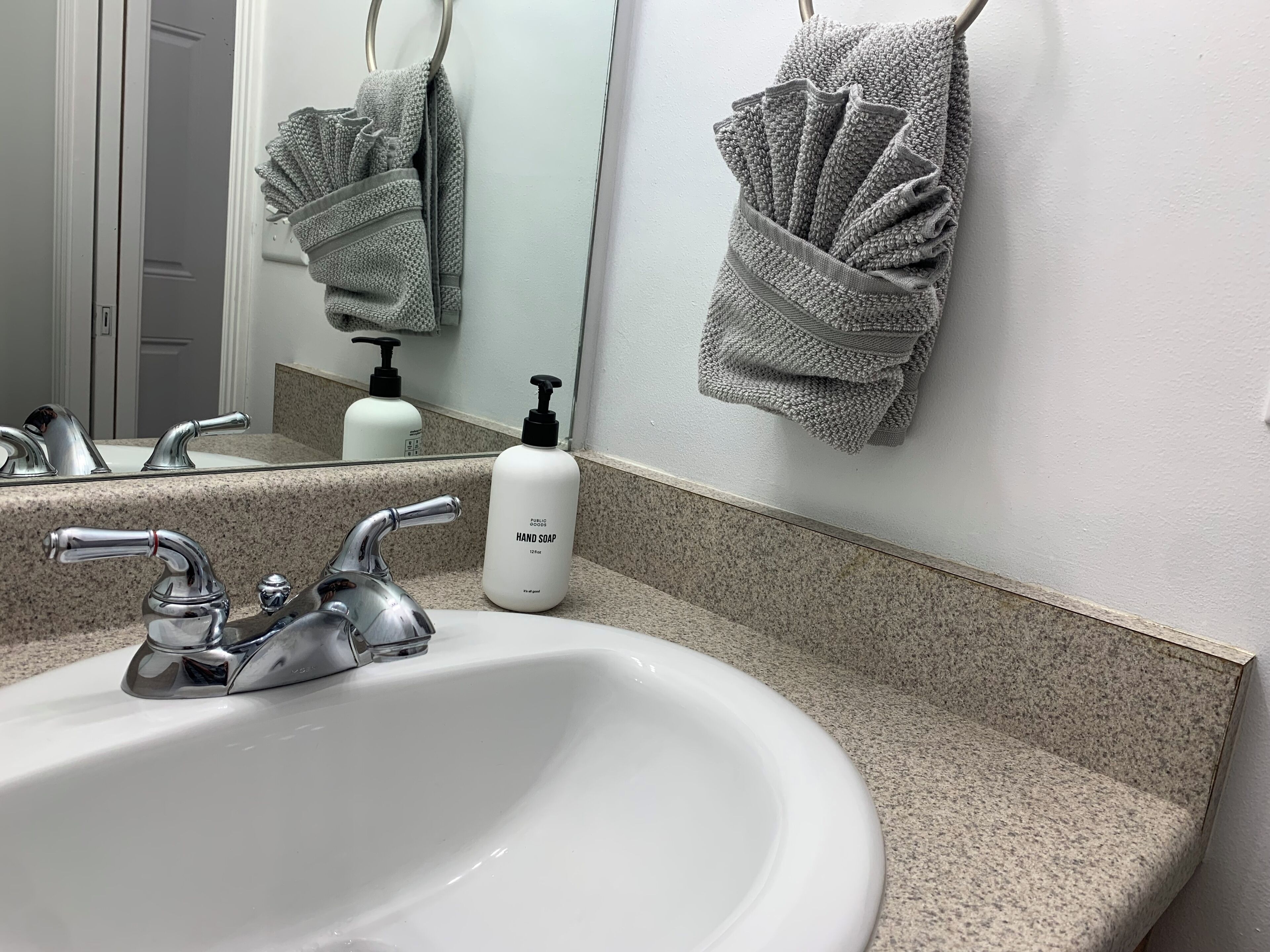Une salle de bain avec un lavabo blanc, deux robinets, un miroir, un porte-serviettes et un flacon de savon pour les mains.