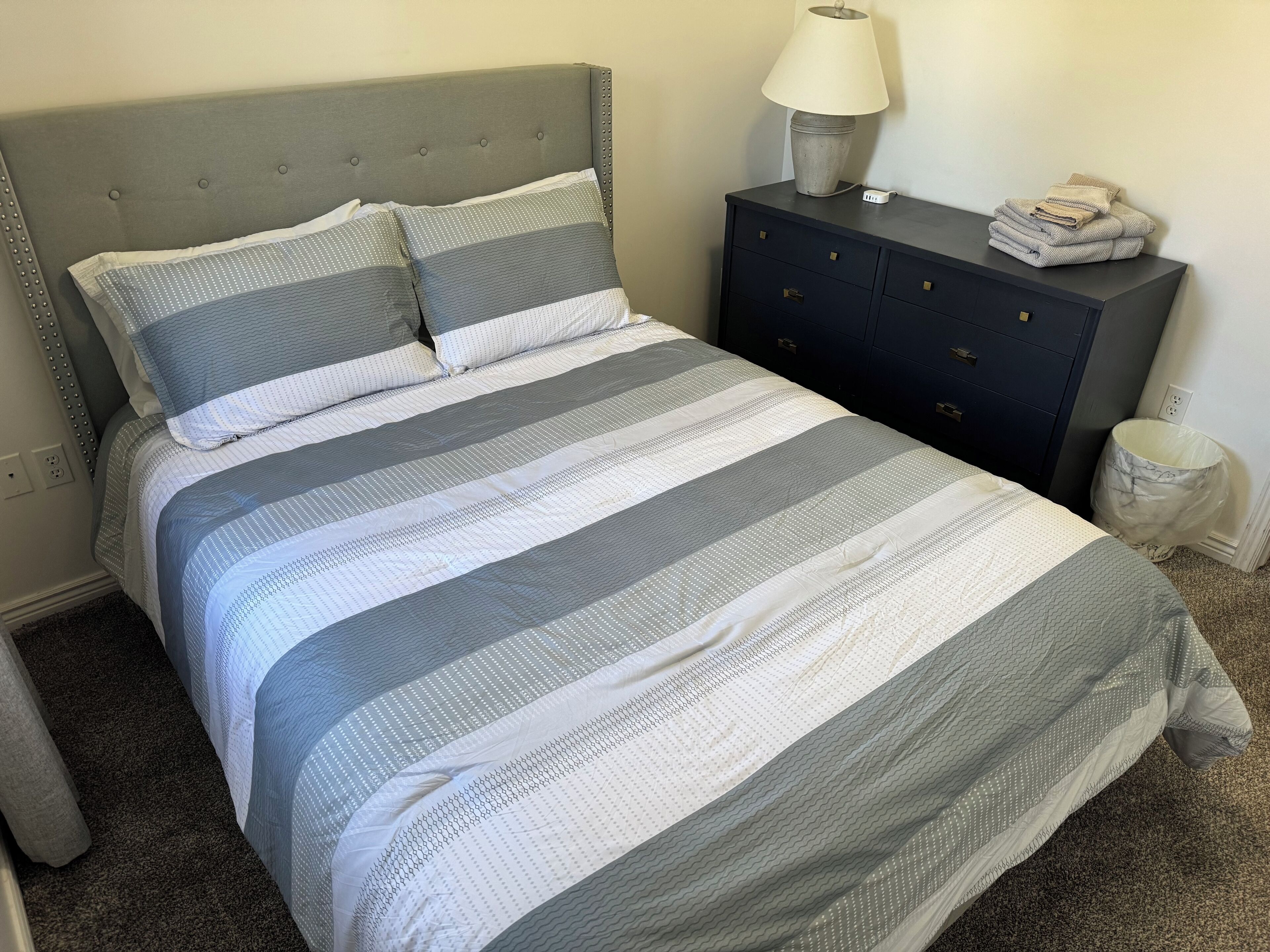 A neatly made bed with a striped bedspread, a grey upholstered headboard, a bedside table with a lamp, and a small trash bin.